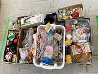 Overview of multiple boxes containing vintage sewing notions including buttons, threads, pins, ribbons, and a battery-operated handheld sewing machine.