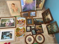 Overview of assorted wall art including framed prints, decorative plates, and embroidery on a tiled floor near a blue wall.