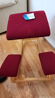Front angle view of ergonomic kneeler chair showing padded seat and knee rests in dark red fabric with natural wood rocker frame.