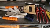 Photo including hand saw, metal file, pruning shears, and white leather gloves, arranged on metal shelf.