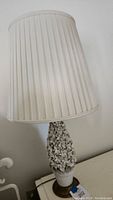 Full view of the lamp showing the white pleated lampshade and ceramic flower pot base with blooming flowers.