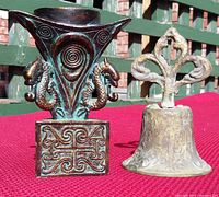 Photo showing both the antique brass church ringer bell with a detailed handle and the decorative candle holder with seahorse designs on a red cloth background.