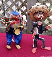 Front view showing two marionette puppets suspended by strings; mariachi on right with sombrero and skeleton suit, clown on left with colorful clothes and drum.