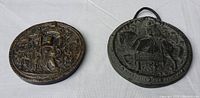 Two round antique reproduction plaques shown side by side on a cloth background. One is a darker, smoother plaque presumably the seal of King James, the other is larger with a rope hanging, the seal of Queen Elizabeth.