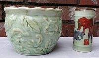 Two celadon crackle glazed pots displayed against a brick wall. Larger pot shows raised botanical relief design, smaller pot shows hand-painted horses.