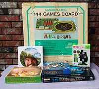 Photo of vintage Carom 144 Games Board box with image of the game and its features, along with five other boxed games and a circular container with a man's face. Shows lot contents clearly.