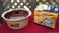 Photo of beige Amana stoneware cooker with glass lid next to boxed London Drugs pasta maker on red surface outdoors.