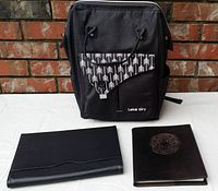 Front view of black travelers backpack with front pocket showing white tree pattern flap and silver clasp, black leather laptop cover, and brown leather notebook with embossed design.