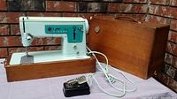 Full view of the vintage soft blue Singer sewing machine on wooden base, with attached cords and foot pedal placed in front, alongside wooden carrying case.