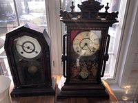 Two vintage wind-up mantel clocks placed side-by-side, one smaller with simpler peaked top and the larger featuring ornate carved top, both showing Roman numeral clock faces, glass front, and pendulums.