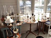 Multiple oil lamps and glass chimneys arranged on small round and side tables in a bright sunroom, seen from multiple angles.