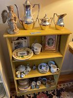 Overview of yellow shelves containing the silverplate tea/coffee set on top, porcelain plates, mugs, creamers, sugars, small dishes on shelves, framed prints, and ceramic angel statue.