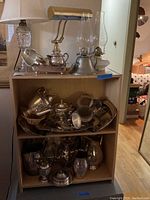 Shelf unit displaying tarnished silver-tone tea set, serving pitchers, and various metalware items along with two oil lamps and a table lamp on top shelf.