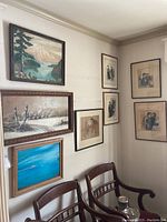 Wall displaying 9 framed pieces including 3 canvas paintings showing landscapes, birds, and ocean scene above wooden chairs.