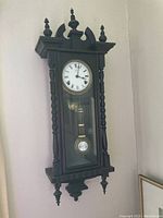 Full view of vintage pendulum wall clock mounted on wall showing entire wooden case, glass front door, and pendulum.
