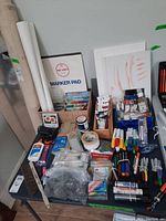 Wide view of art supplies on table showing marker pad, canvases, glue, tapes, brushes, paints, markers, and clays.