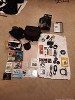 Overview of entire lot on beige carpet: black camera bag center, Sony telephoto zoom lens in cloth pouch at left, assorted manuals and software CDs at bottom, filter boxes and cables at right