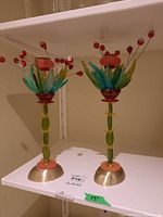 Pair of colorful decorative candle sticks with glass floral and berry elements and metal bases, shown on white shelf.