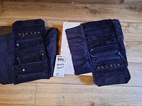 Two navy blue fabric jewellery pouches laid open on wooden floor showing multiple compartments, straps with snaps, and eyelets for jewellery storage.