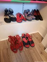 Photo showing seven pairs of ladies shoes arranged on a shelf and floor including black strappy heels, red clogs, multicolor patterned heels, black dress heels, and red sandals.