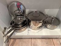 Set of 10 stainless steel pots and pans including saucepans, stockpot, and lids stored in a cabinet shelf.