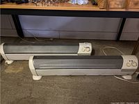 Two white electric space heaters with metal mesh heating surfaces placed on floor; control panels visible on side with adjustment knobs and indicator lights.