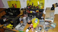 Wide view of two open plastic toolboxes containing various hand tools, work boots, tape rolls, and hardware items spread on a table.
