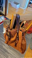 Fossil brown leather tote bag hanging on a wooden chair, showing front and side view with straps and buckle details.