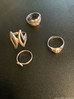 Four silver rings laid on black surface showing various designs including a cat face with missing eye stone and a geometric ring