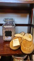 Pair of tan suede leather slippers with cream shearling lining next to a large glass jar filled with dried lavender flower tops on a wooden shelf.