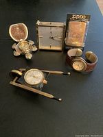 Wide shot of all items on black surface: turtle watch, Cyma desk clock, wheelbarrow watch, wristwatch, boxed Zippo lighter.