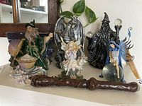 Front view of two wizard figurines, two fairy figurines, a dragon figurine, and a wooden wand on a shelf with houseplant background.