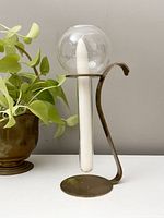 Brass and glass candlestick holder with a curved handle, round base, and clear glass globe around the candle area, placed beside a plant.
