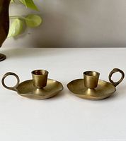 Pair of small brass chamber candlestick holders shown side by side on white surface with plant in background.