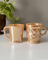 Two pink vintage Anchor Hocking mugs placed side by side on a white surface against a gray background with a plant in the background.