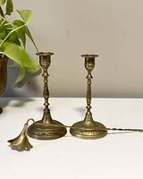 Front view of both brass candlestick holders on table with candle snuffer laid across base showing engraved floral patterns and patina.