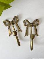 Two brass ribbon motif candlestick holders displayed on white surface showing front view, highlighting the bow design and candle cup.