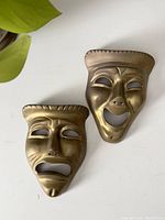 Front view of two brass theatre masks, comedy and tragedy, on a white surface with nearby green leaf.