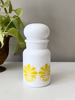 Front view of white opaque jar with yellow floral decoration and bubble top lid, sitting on a white surface with plant in background.