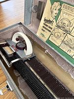 Close-up of vintage manual knitting machine inside case showing white handle, red dials, and needle bed. Instruction manual attached inside case lid.