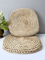 Two large square coiled wicker seating cushions placed on a white table against a light grey wall with some green plant leaves visible to the side.