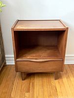 Full front view of wooden bedside table on hardwood floor showing open shelf, drawer, and splayed legs