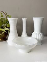 Three vintage milk glass vases and one candy dish displayed on a white surface against a light gray wall, styled next to a green potted plant.