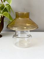 Front view of the vintage mushroom candle jar showing amber top and clear cylindrical base on white surface with a plant beside it.
