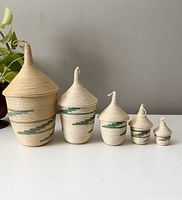 Five vintage nesting coiled baskets with pointed lids displayed side by side from largest to smallest. Beige and green woven material with geometric pattern.