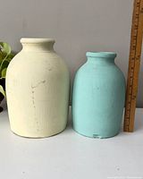 Front view of two vintage crocks, cream and light blue, placed side by side with a measuring stick showing heights.