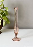 Full frontal photo of a tall, slender pink glass vase with a round base on a white surface, green plant in background.