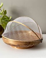 Side angle of the round wicker picnic basket with white mesh food cover raised partially, showing woven basket detail and mesh dome shape frame.