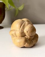 Photo of the small wooden Buddha statue showing its curled posture with head bowed, placed on white surface with green plant matter in background.