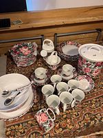 Full view of the dinnerware set laid out on a patterned fabric surface including plates, bowls, cups, and accessories with the signature rose garden floral pattern.